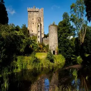Historic Blarney Castle with stone towers, surrounded by greenery and a moat, under a clear blue sky in Ireland.