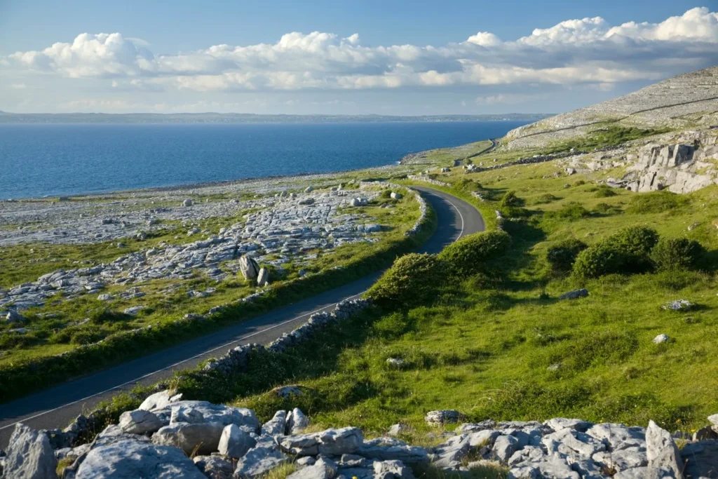 Driving through the Burren: a winding road cuts through the unique limestone landscape, with rocky terrain and green patches leading to the stunning blue ocean under a partly cloudy sky.