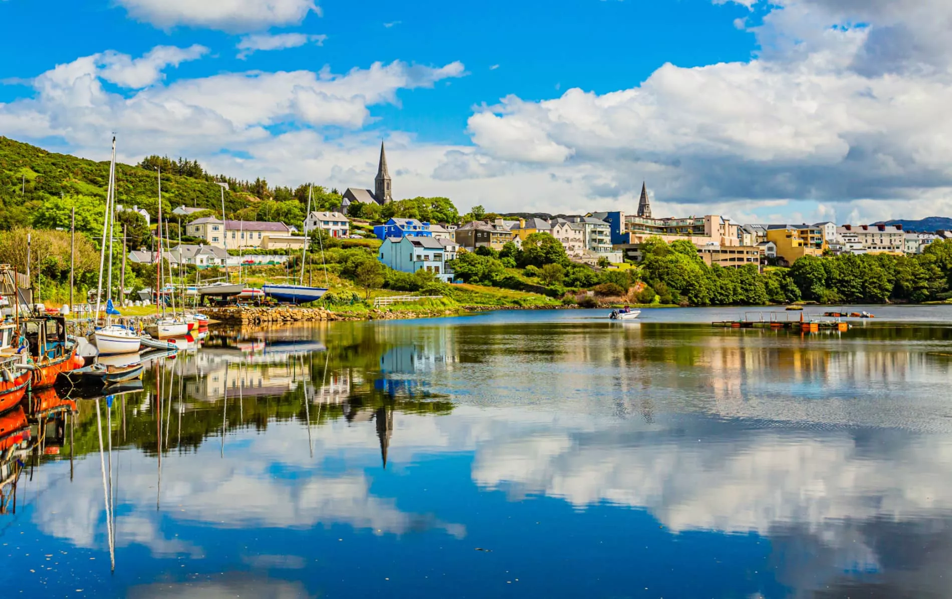 View of colorful waterfront buildings, boats, and church spires reflected in the calm waters of Clifden, a charming coastal town in Galway, Ireland – part of the 2-day Galway and Connemara tour with Irish Chauffeur Transfers.
