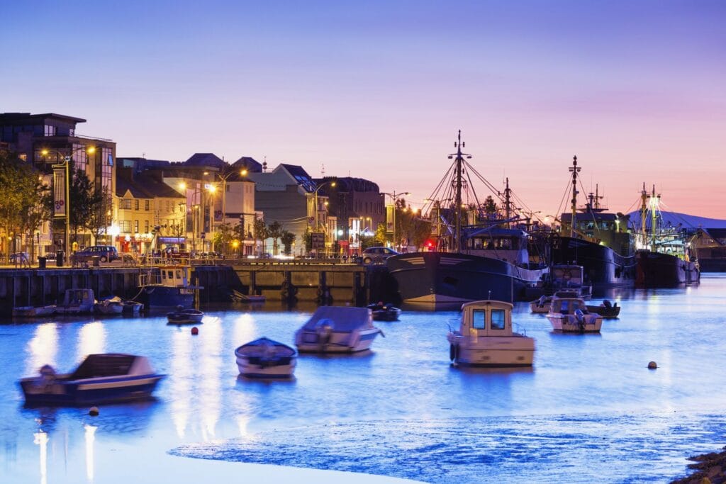 Evening view of Waterford Harbour with waterfront buildings and boats under a darkening sky.