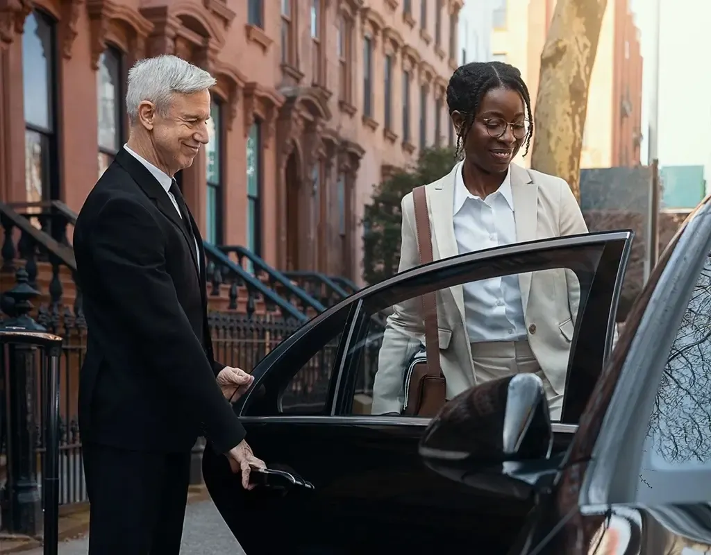 A driver from Chauffeur Service company in Dublin, in a black suit opens a limousine door for a passenger in a city street with brownstone buildings.